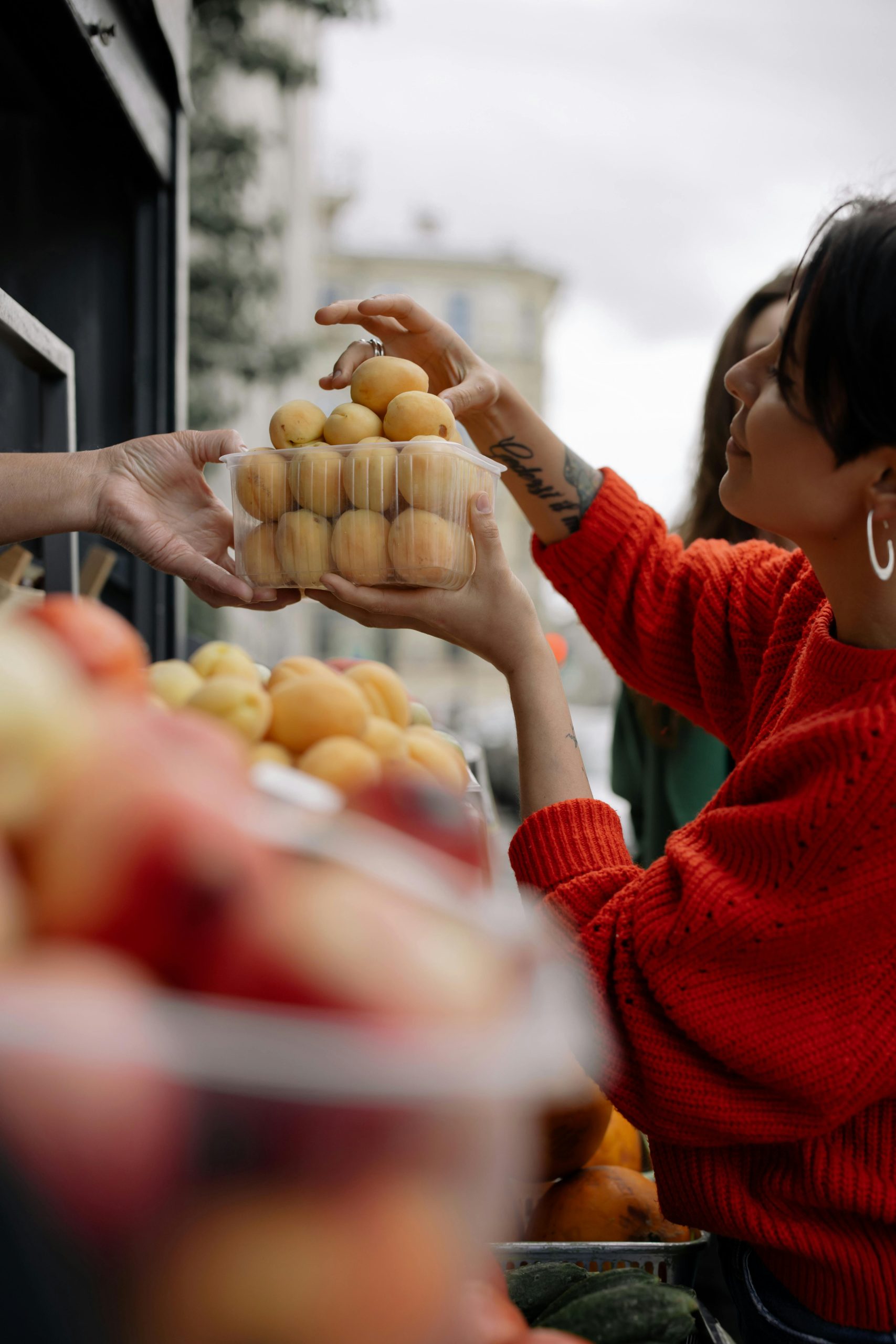 A woman in a red sweater purchases apricots at an outdoor fruit market stall.